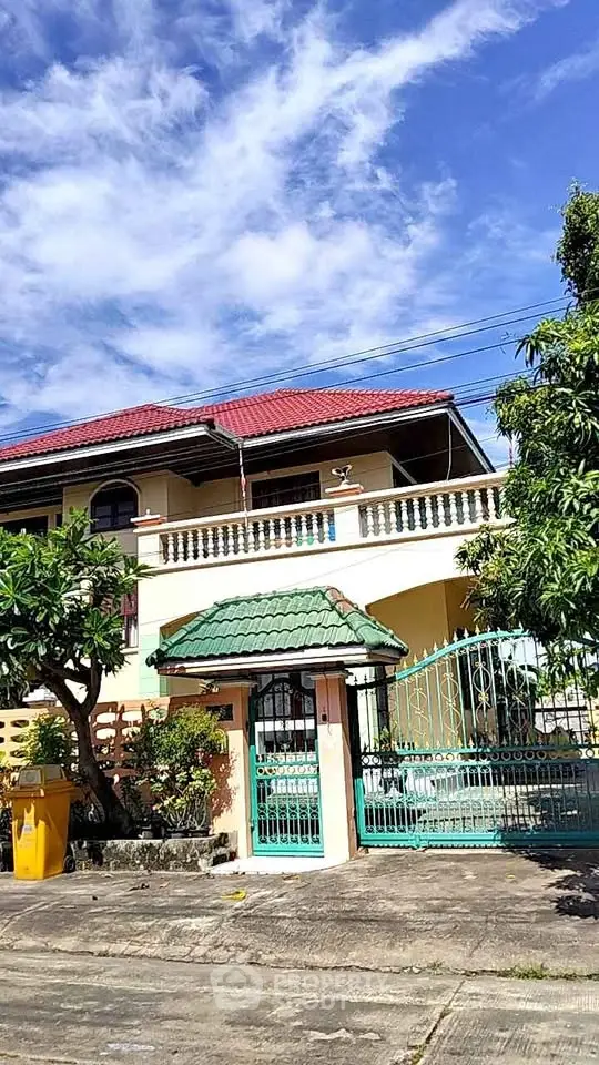 Charming two-story house with red roof and green gate, surrounded by lush greenery and clear blue skies.