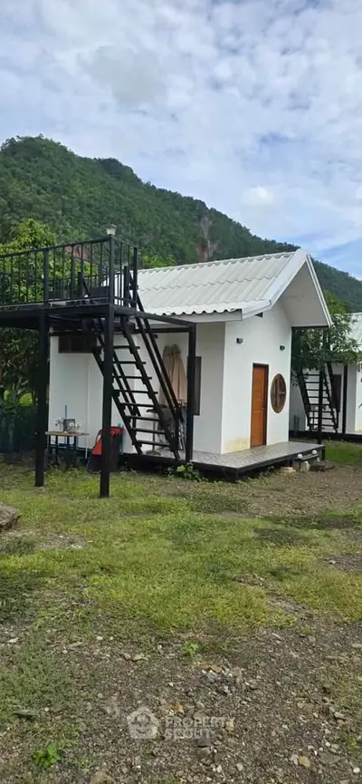 Charming small white house with rooftop terrace set against lush green mountain backdrop.