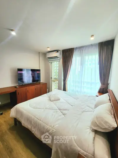 Spacious bedroom with modern decor, featuring a large bed, TV, and natural light from a balcony window.