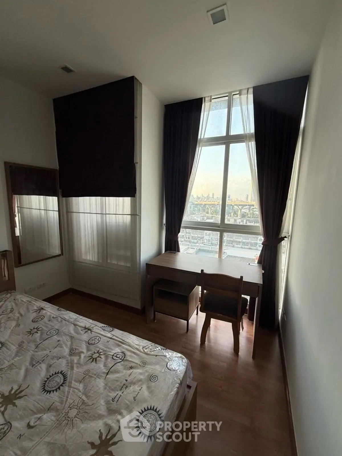 Cozy bedroom with large window offering stunning city view, featuring a wooden desk and chair.