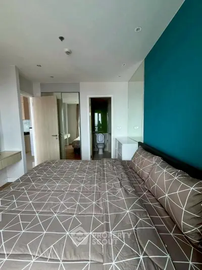 Modern bedroom with geometric bedding and teal accent wall, featuring a view into an ensuite bathroom.