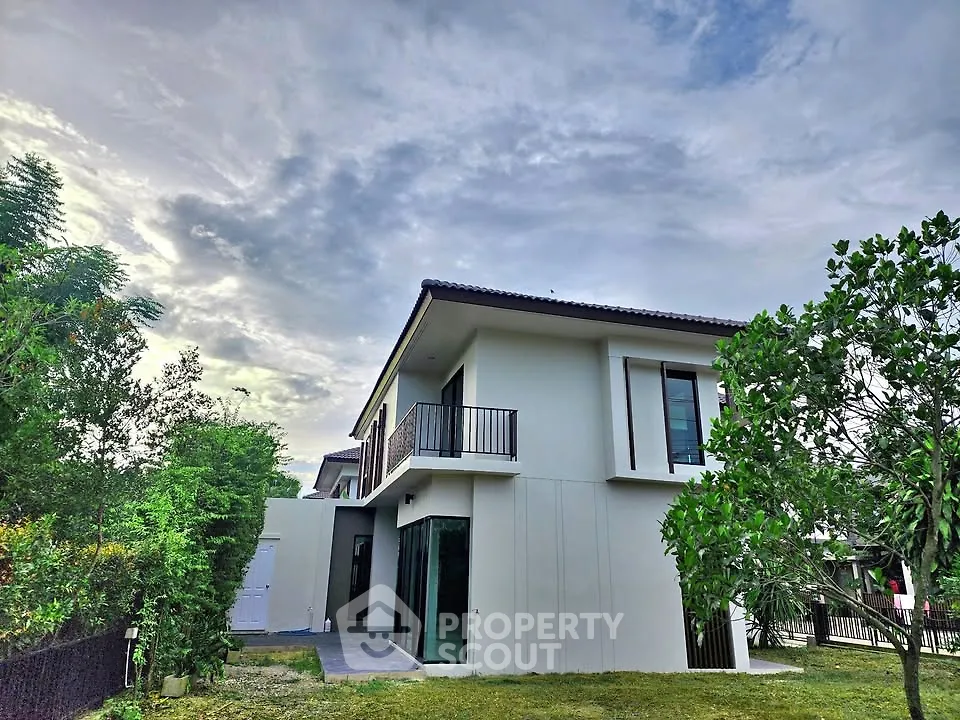Modern two-story house with lush garden and balcony under cloudy sky.