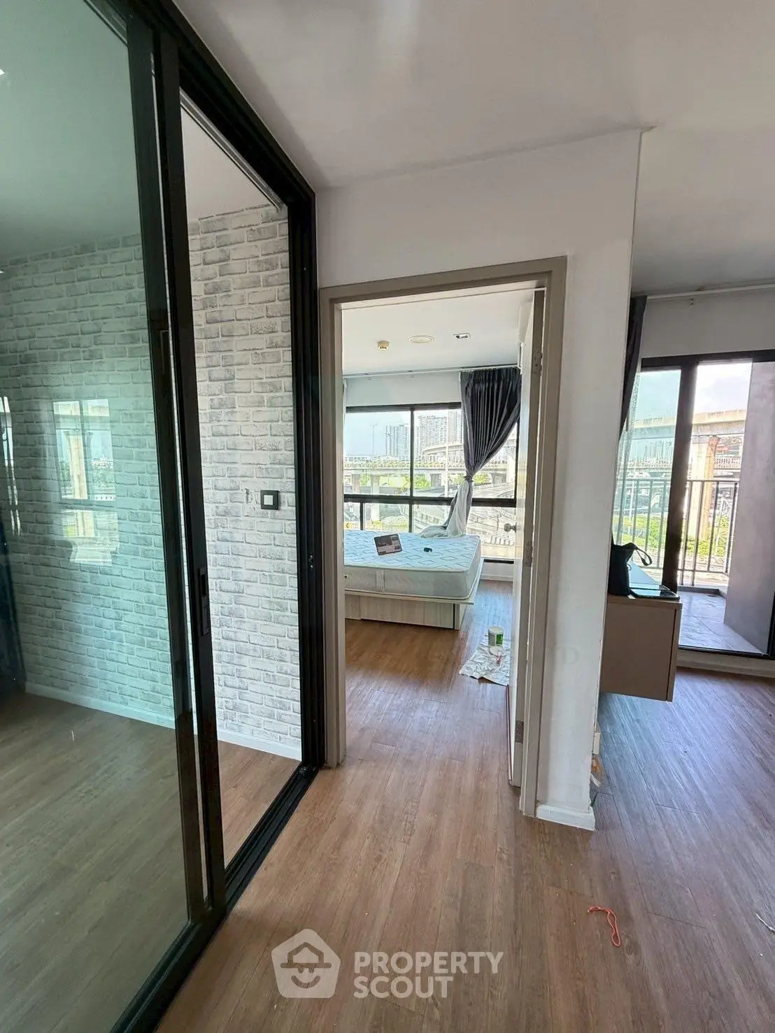 Modern apartment hallway leading to a bright bedroom with balcony view, featuring stylish wood flooring and brick accent wall.