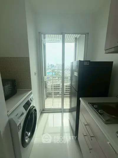 Modern kitchen with city view, featuring washing machine, fridge, and microwave, leading to a balcony.