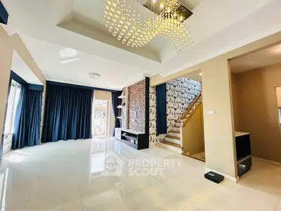 Spacious modern living room with elegant chandelier and staircase, featuring large windows and stylish decor.