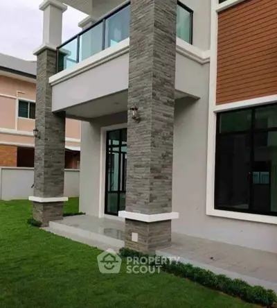Modern two-story house exterior with stone accents and glass balcony, surrounded by lush green lawn.