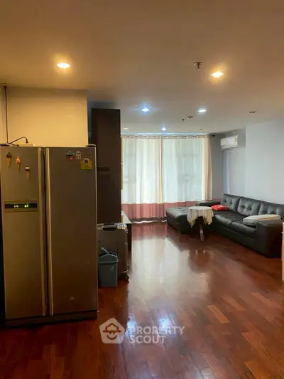 Spacious living room with wooden flooring and modern furniture, featuring a large fridge and air conditioning.