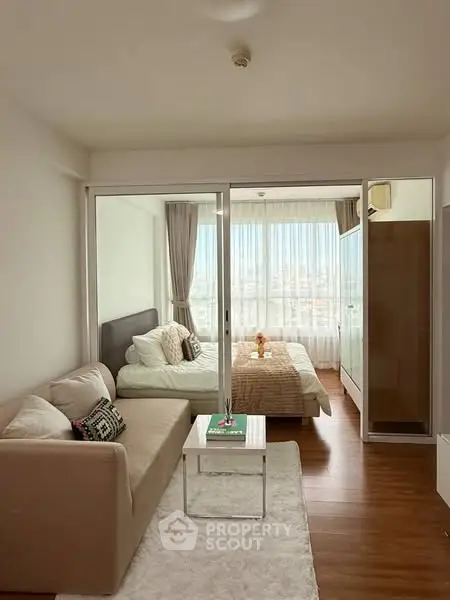 Cozy studio apartment with bed and sofa, featuring large windows and wooden flooring.