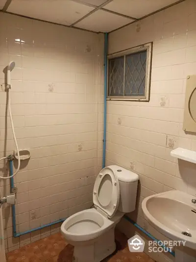 Compact bathroom with full tiling, shower area, toilet, and pedestal sink, featuring a small diamond-patterned window for natural light.