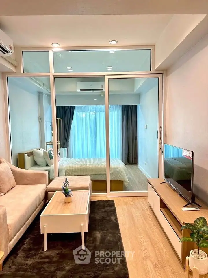 Modern living room with glass partition leading to a cozy bedroom, featuring stylish furniture and natural light.