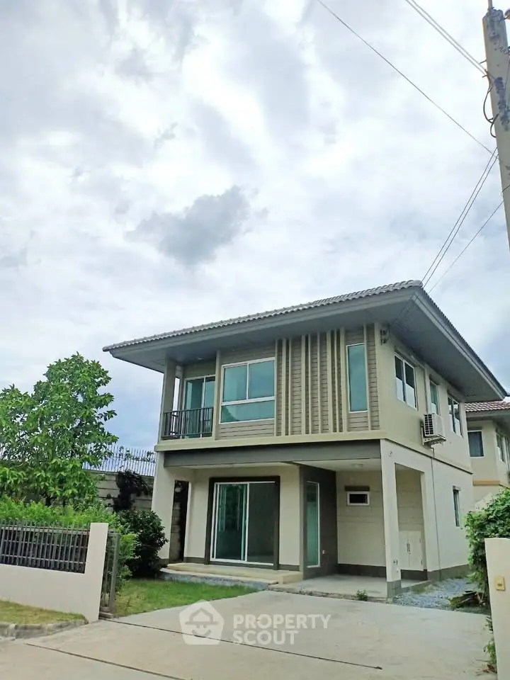 Modern two-story house with large windows and a small balcony, surrounded by greenery and a paved driveway.