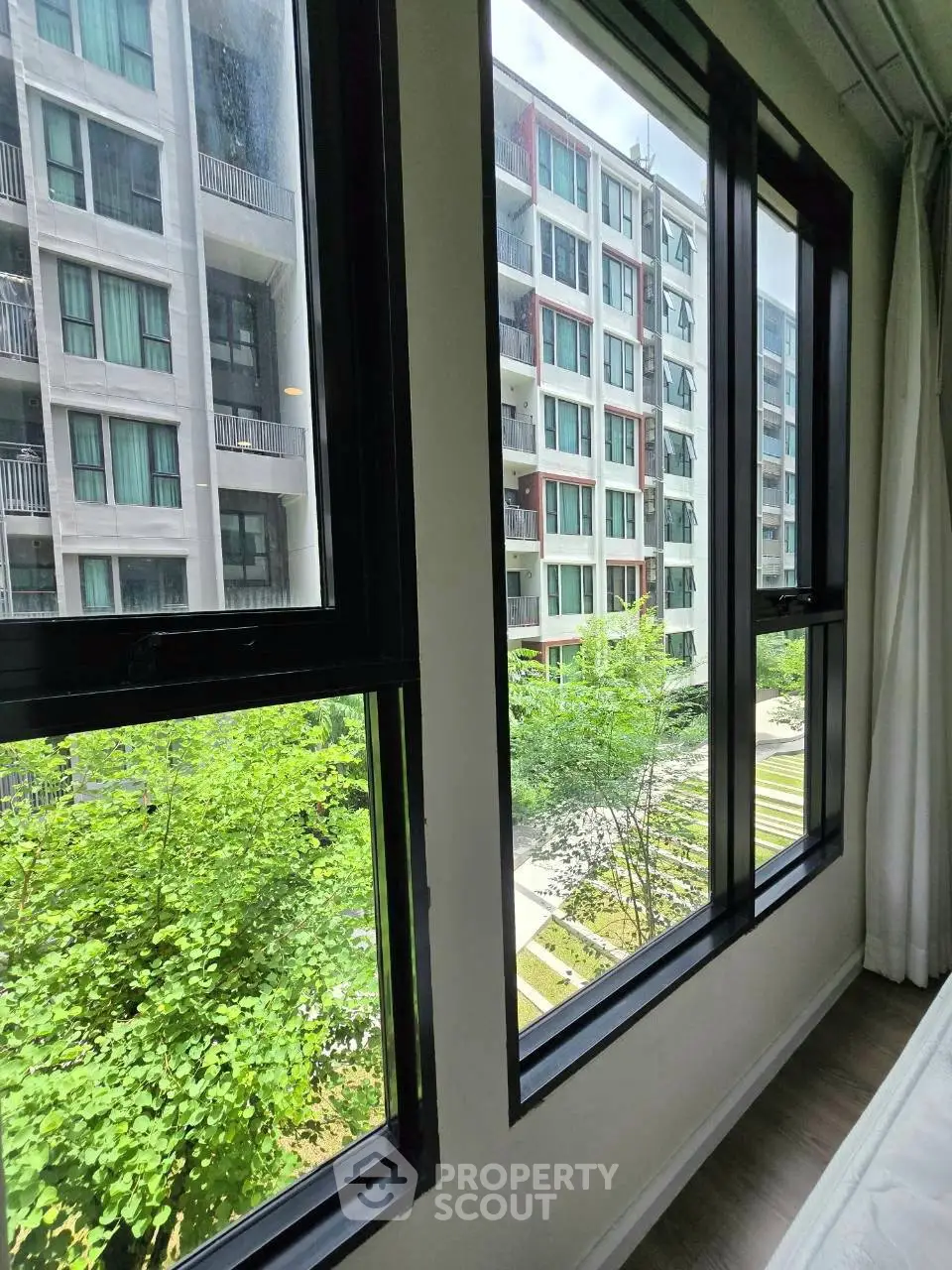 Modern apartment view with large windows overlooking lush greenery and contemporary buildings.