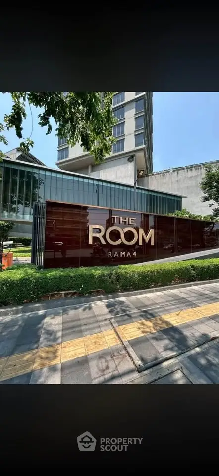 Modern condominium entrance with sleek design and lush greenery at The Room Rama 1.