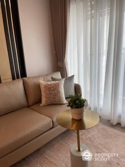 Elegant living room with beige sofa, stylish cushion, and gold accent table near sheer curtains.