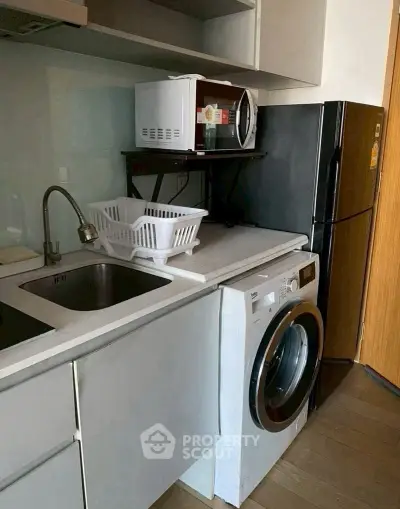 Compact kitchen with washing machine, microwave, and fridge in modern apartment.