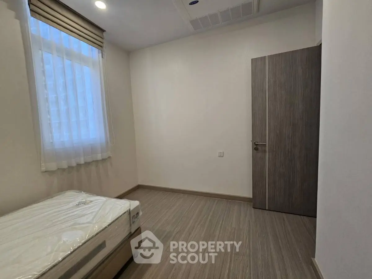 Minimalist bedroom with wooden flooring and natural light from window.