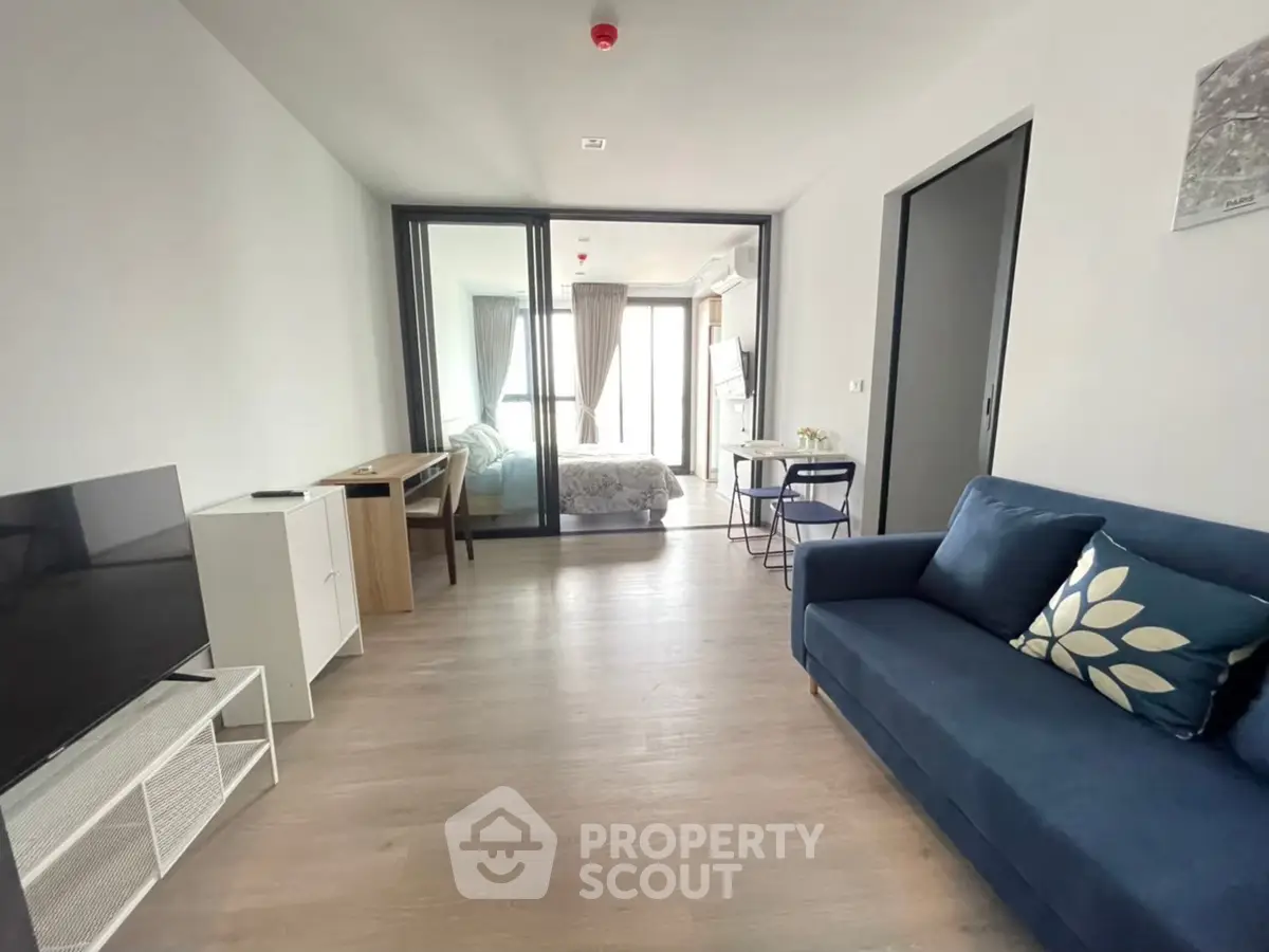 Modern living room with blue sofa and TV, leading to a cozy bedroom with natural light.
