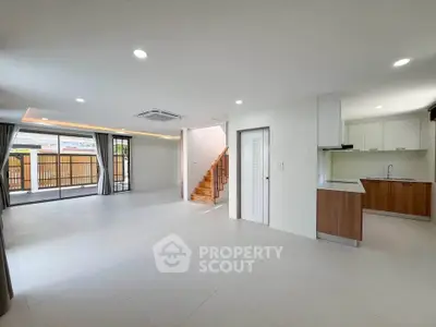 Spacious open-plan living room with modern kitchen and staircase, featuring large windows and natural light.