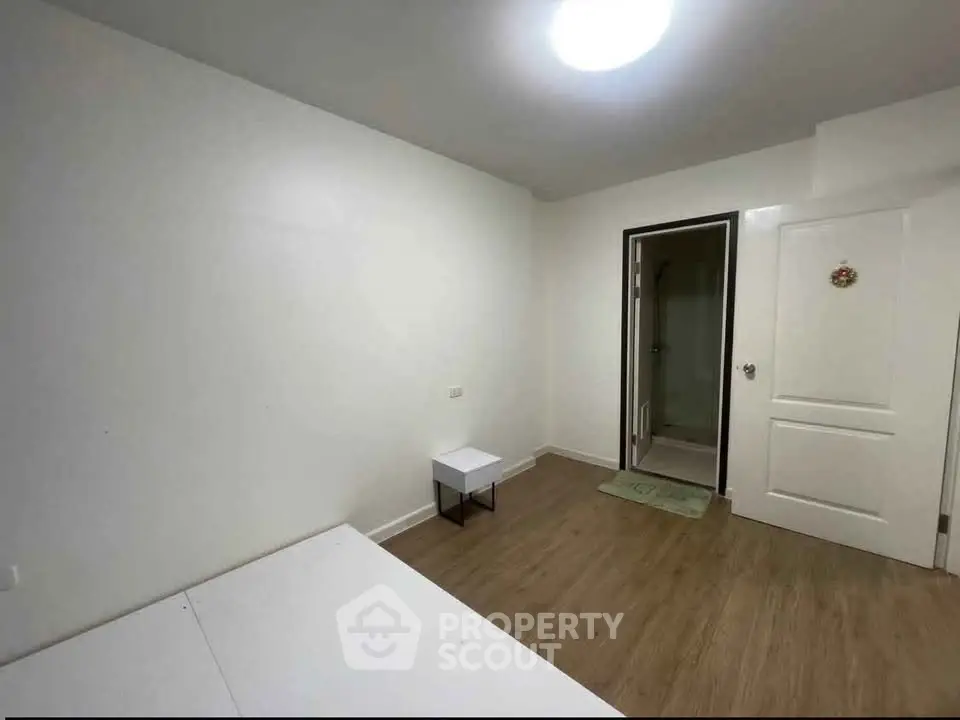 Minimalist bedroom with wooden flooring and white walls, featuring a small table and a door leading to an ensuite bathroom.