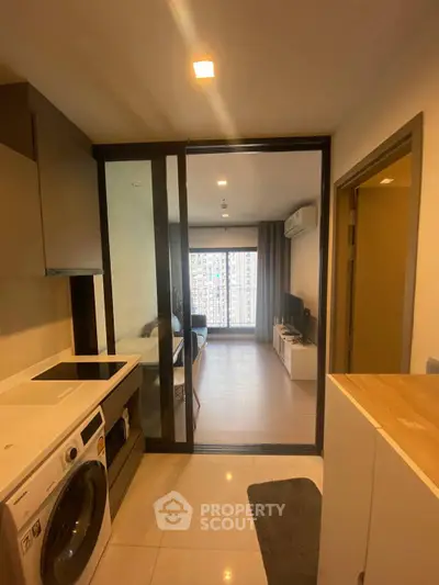 Modern apartment interior with open kitchen and living room, featuring washing machine and large window view.