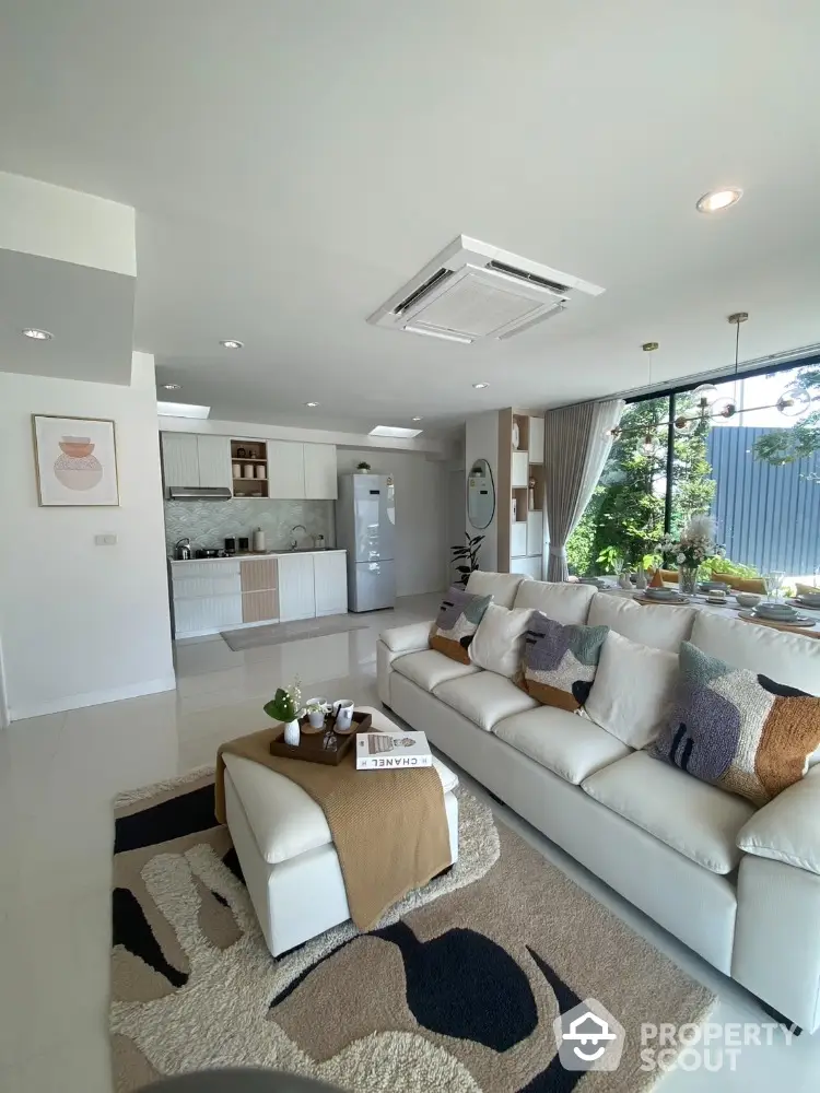 Modern living room with open kitchen, stylish decor, and large windows overlooking greenery.