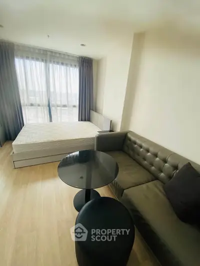Modern studio apartment with bed, sofa, and large window offering natural light.