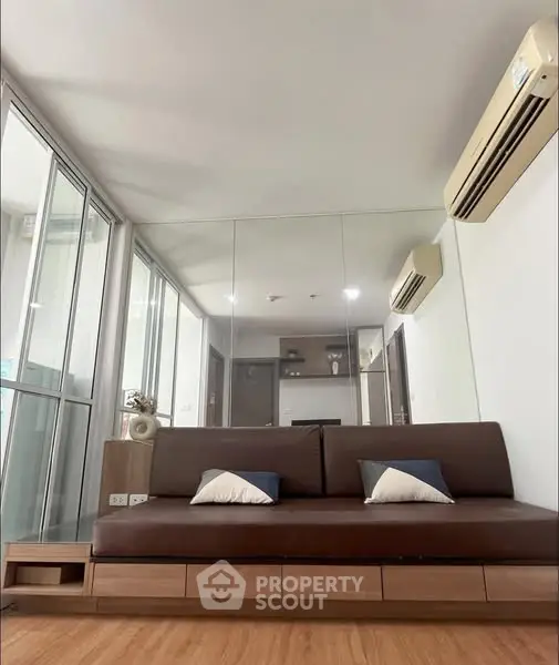 Modern living room with sleek brown sofa and mirrored wall, featuring air conditioning and wooden flooring.