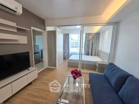 Modern living room with glass partition to bedroom, featuring sleek furniture and wooden flooring.