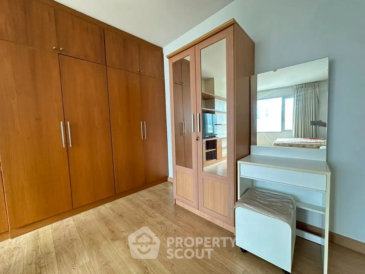 Spacious bedroom with wooden wardrobe and vanity set, featuring large window for natural light.