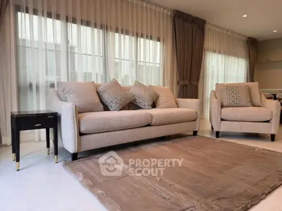 Elegant living room with plush beige sofa and armchair, soft rug, and large windows with sheer curtains.