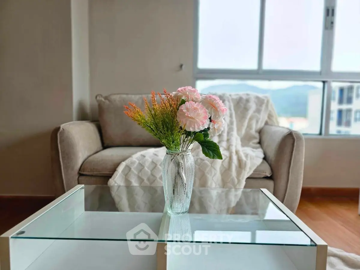 Cozy living room with elegant sofa and glass coffee table, featuring a vase of flowers and a scenic window view.
