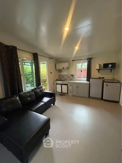 Modern living room with sleek black sofa and compact kitchen area, featuring large windows and stylish decor.
