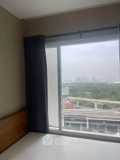 Bedroom with large window offering city and greenery view, modern and cozy ambiance.