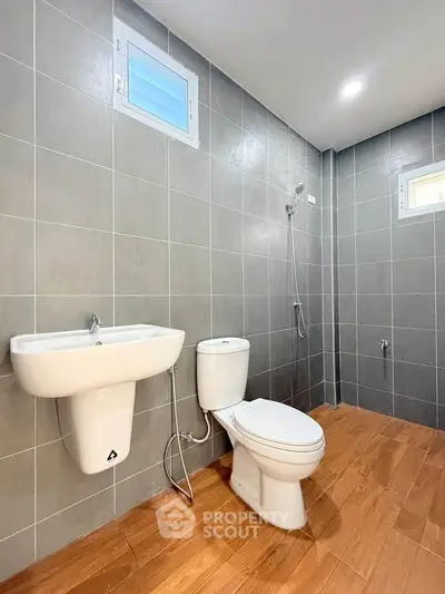 Modern bathroom with sleek gray tiles and wooden flooring, featuring a toilet, sink, and shower area.