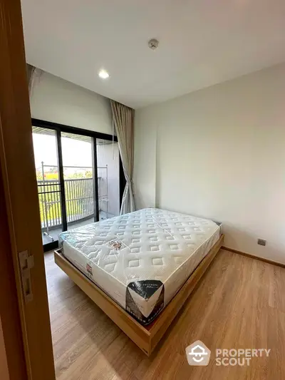 Modern bedroom with large window and balcony access, featuring a comfortable mattress and wooden flooring.