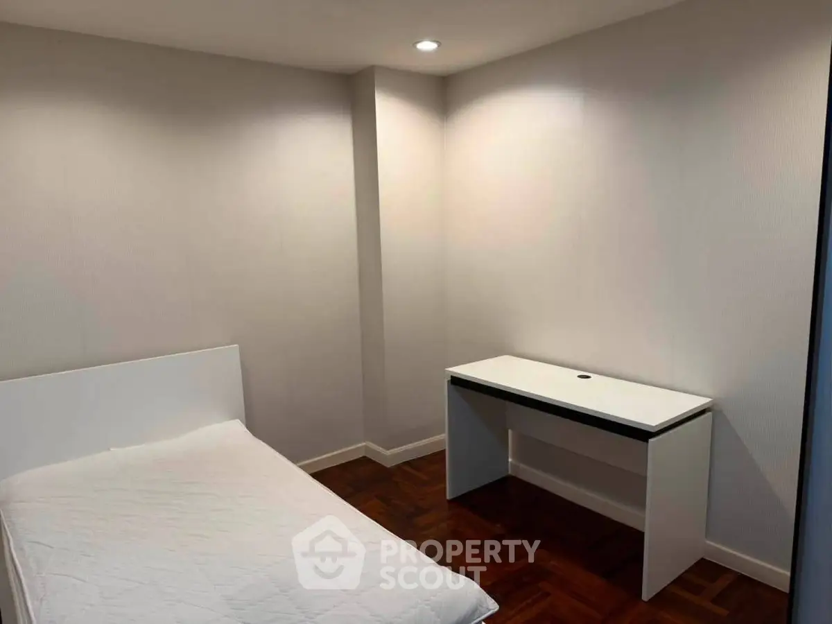 Minimalist bedroom with single bed and desk, featuring wooden flooring and recessed lighting.