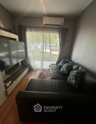 Cozy living room with modern black sofa and large TV, featuring a view of lush greenery through glass doors.