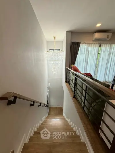 Elegant staircase with chandelier leading to a cozy living room with large windows and modern decor.