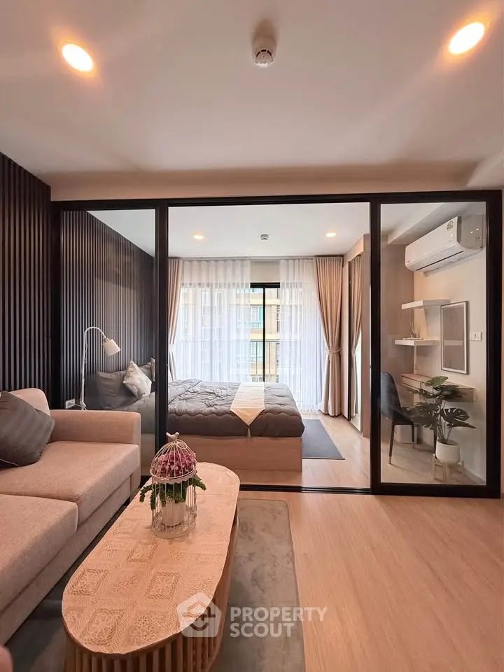 Modern living room with glass partition leading to a cozy bedroom, featuring stylish decor and natural light.