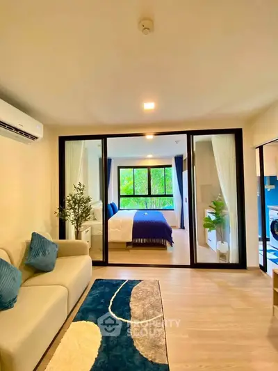 Modern open-plan living room with glass partition leading to a bright bedroom, featuring stylish decor and natural light.