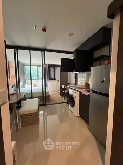 Modern studio apartment with open kitchen and washing machine, featuring large windows and stylish decor.