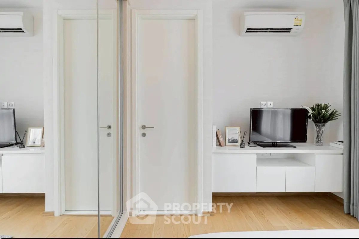 Modern bedroom with mirrored closet and wall-mounted TV, featuring sleek white furniture and air conditioning.
