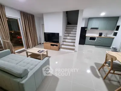 Modern open-plan living room with sleek kitchen and staircase, featuring stylish furniture and large windows.
