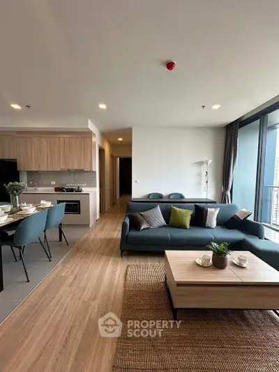 Modern open-plan living room with stylish furniture and kitchen area, featuring large windows and natural light.