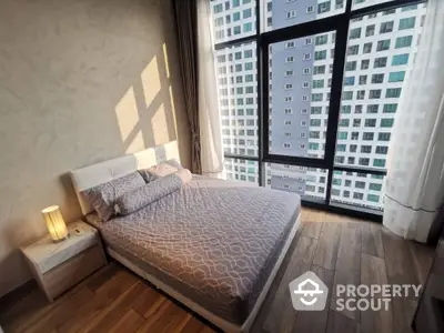 Sunlit bedroom with a large window offering a city view, featuring a cozy double bed and modern wooden flooring.