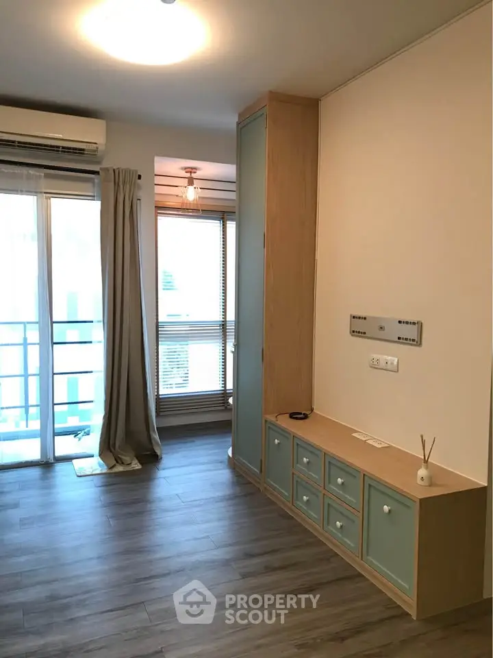 Modern living room with wooden flooring and balcony access, featuring built-in storage and air conditioning.