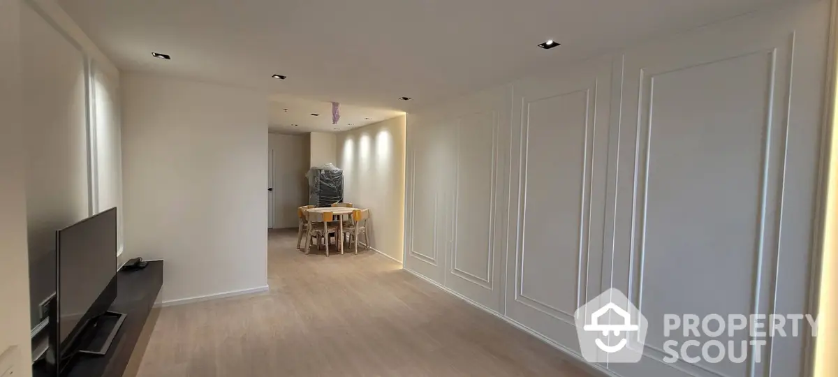 Spacious living room with elegant wall paneling and modern recessed lighting, leading to a cozy dining area with natural light.