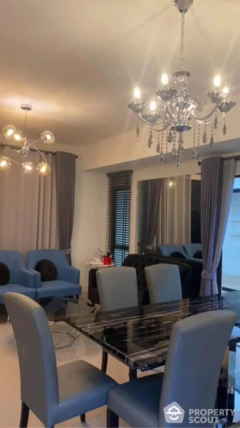 Elegant dining area with modern chandeliers, sleek glass table, and plush blue chairs, complemented by stylish window treatments.