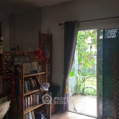 Cozy living room with sliding glass door opening to a lush garden view, featuring a bookshelf and elegant curtains.
