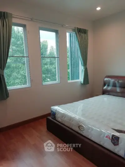Spacious bedroom with large windows and wooden flooring, offering a serene view of greenery.